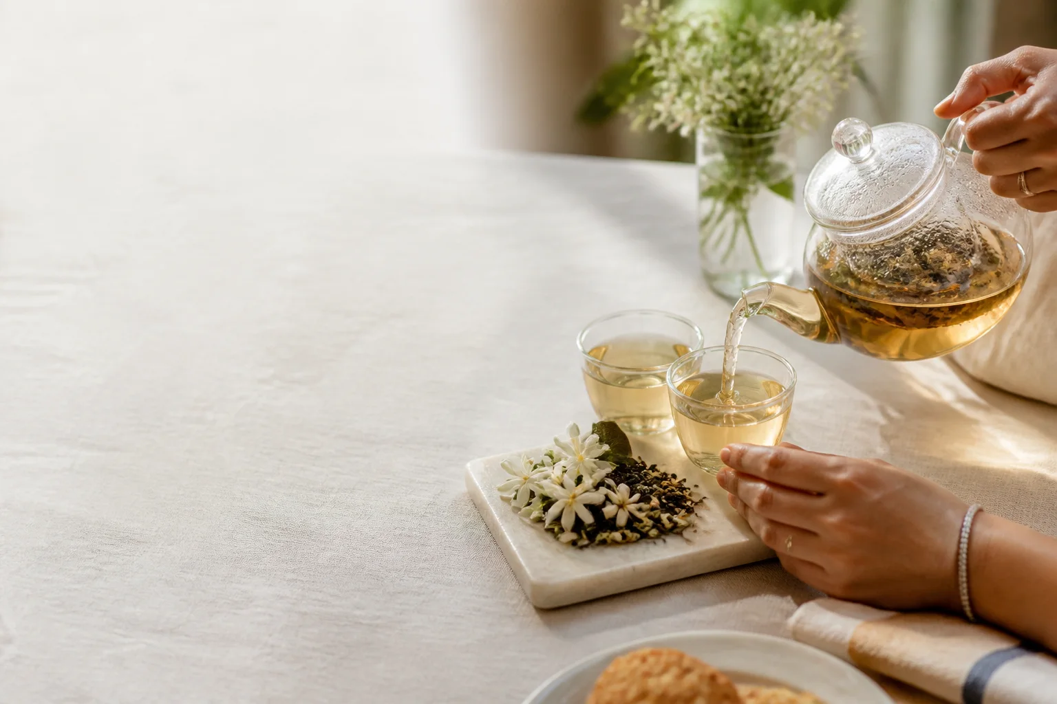 Jasmine blossoms and green tea leaves beside pale golden tea being shared at a sunlit table