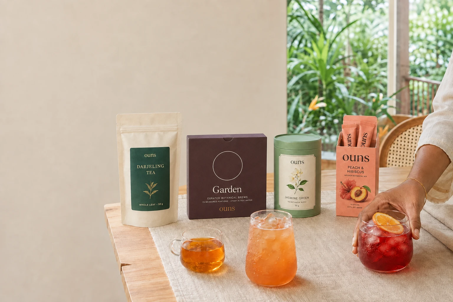 Darjeeling tea pouch, Garden botanical carton, Jasmine Green tea caddy and Peach & Hibiscus instant sticks beside three drinks on a bright breakfast table, with a hand holding the ruby iced infusion